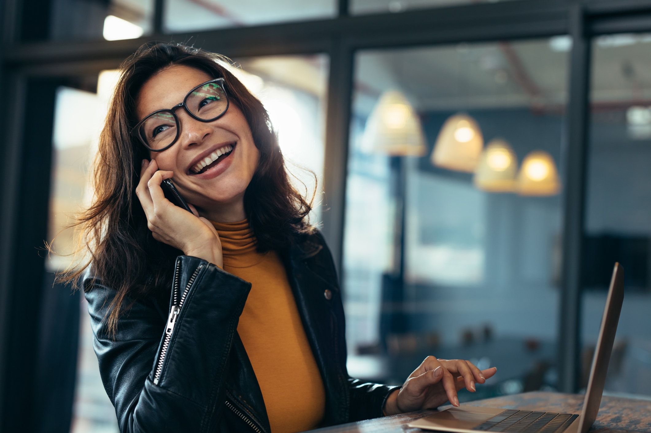 Cheerful asian female talking on mobile phone while sitting on desk with laptop. Business woman in casuals making a phone call and laughing.