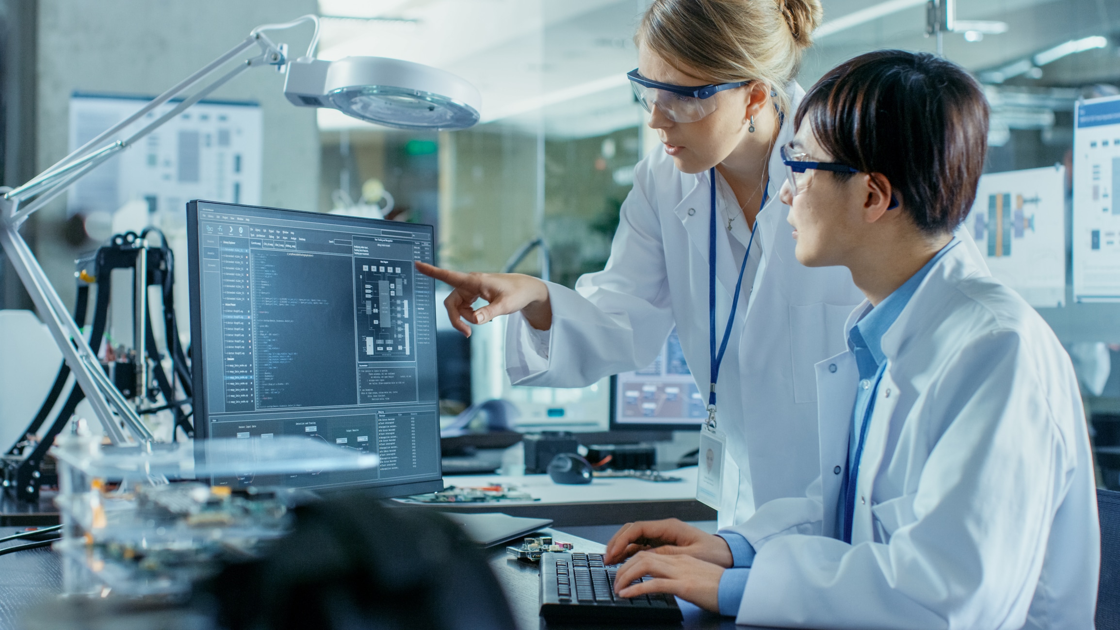 Asian Scientist Sitting at His Desk Consults Senior Engineer about Sophisticated Coding and Programming. In the Background Computer Science Research Laboratory with Robotic Arm Model.