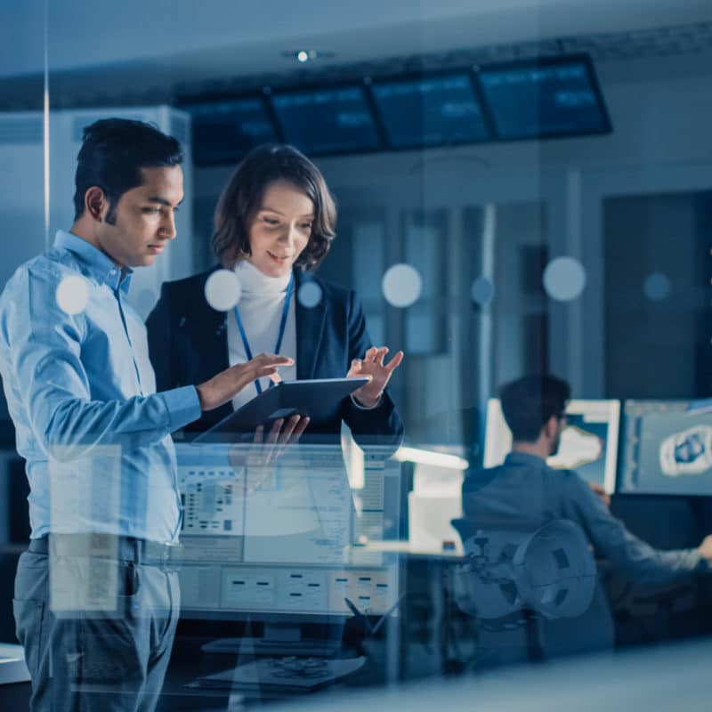 In Technology Research Facility: Female Project Manager Talks With Chief Engineer, they Consult Tablet Computer. Team of Industrial Engineers, Developers Work on Engine Design Using Computers